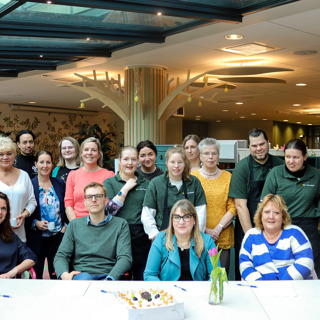 Grote groep mensen zit aan tafel met taart voor het tekenmoment van de  samenwerkingsovereenkomst voor de huiskamer voor de buurt