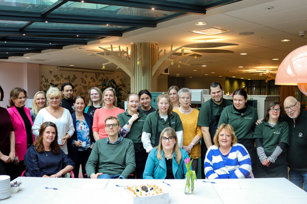 Grote groep mensen zit aan tafel met taart voor het tekenmoment van de  samenwerkingsovereenkomst voor de huiskamer voor de buurt